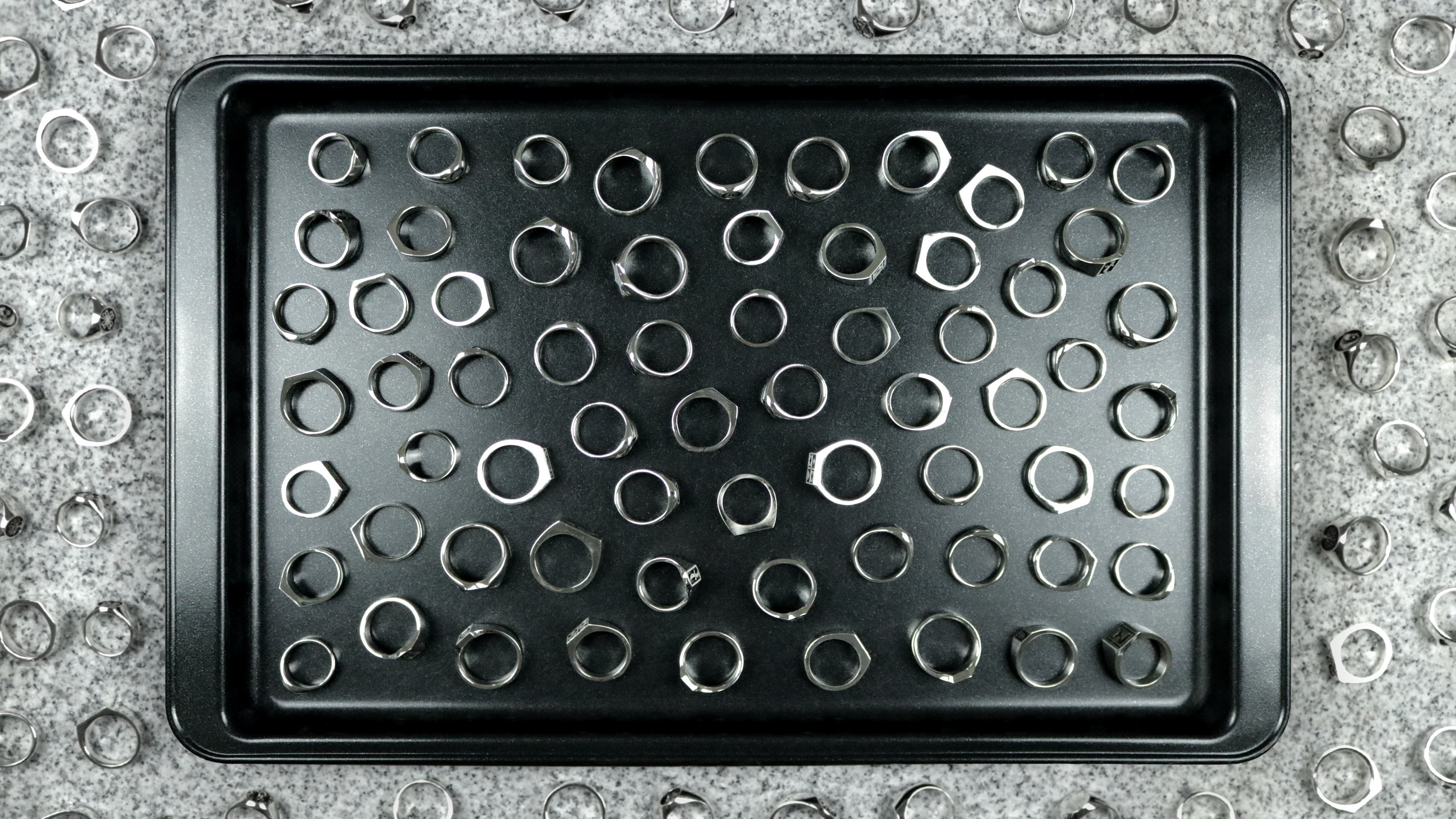 Overhead view of Jack Sterling sterling silver signet rings arranged in a black tray, surrounded by additional rings, showing scale of the collection
